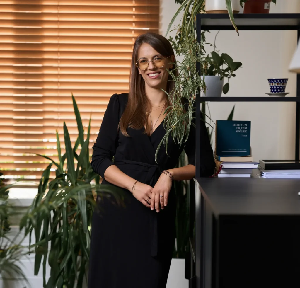 Martyna Ludwiczak w czarnym stroju stojąca w kancelarii na tle regału z książkami i roślinami